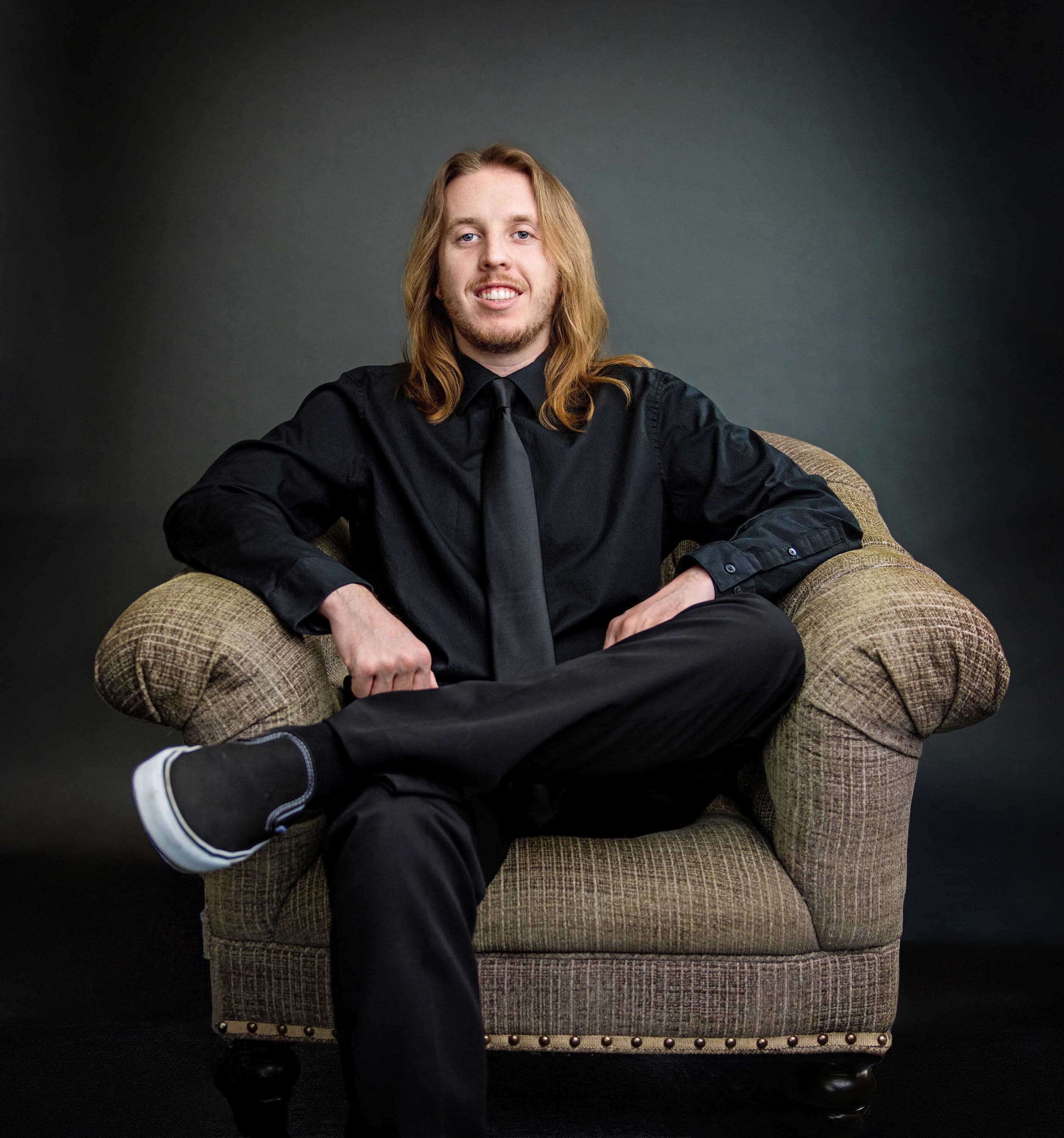 Studio headshot of a professional in a black shirt and tie seated in an upholstered armchair against a controlled dark gray backdrop