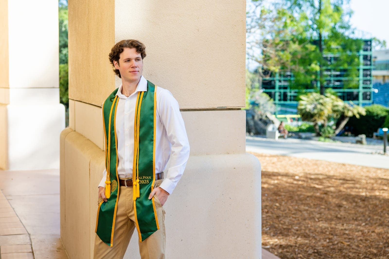 Senior in a white button-down and graduation stole leaning against a stucco column on a sunlit campus during a Central Coast senior portrait session