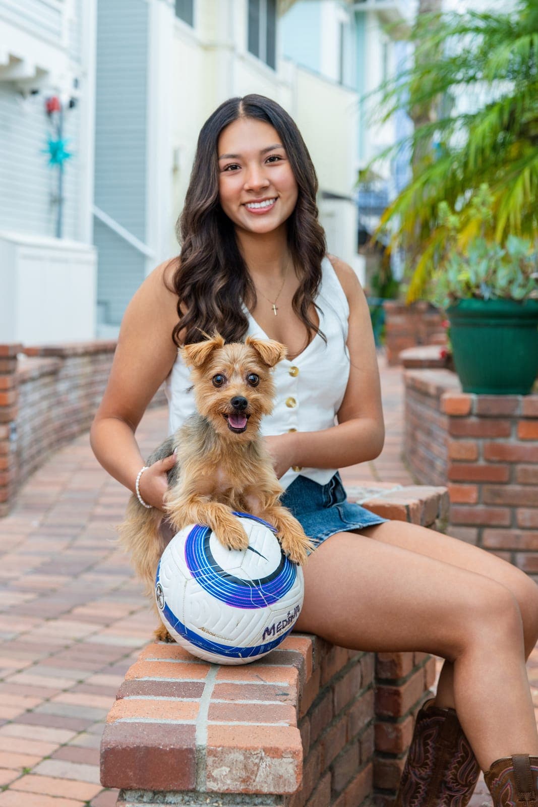 High school senior in a white vest and denim skirt sitting on a brick wall holding her small terrier, with her soccer ball resting against the bricks in front of her